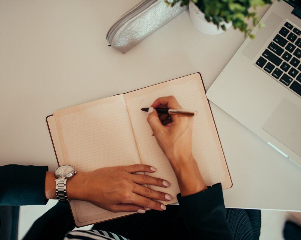 يد تكتب في دفتر مفتوح بجوار حاسوب محمول على مكتب أبيض